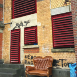 Ein braunes Ledersofa steht auf einem Bürgersteig vor einem alten Backsteingebäude mit roten Fensterläden, Graffiti und einer blauen Plastiktüte. In der rechten oberen Ecke steht Werkstatt_ und_Freiheit und Rheinischer Verein.